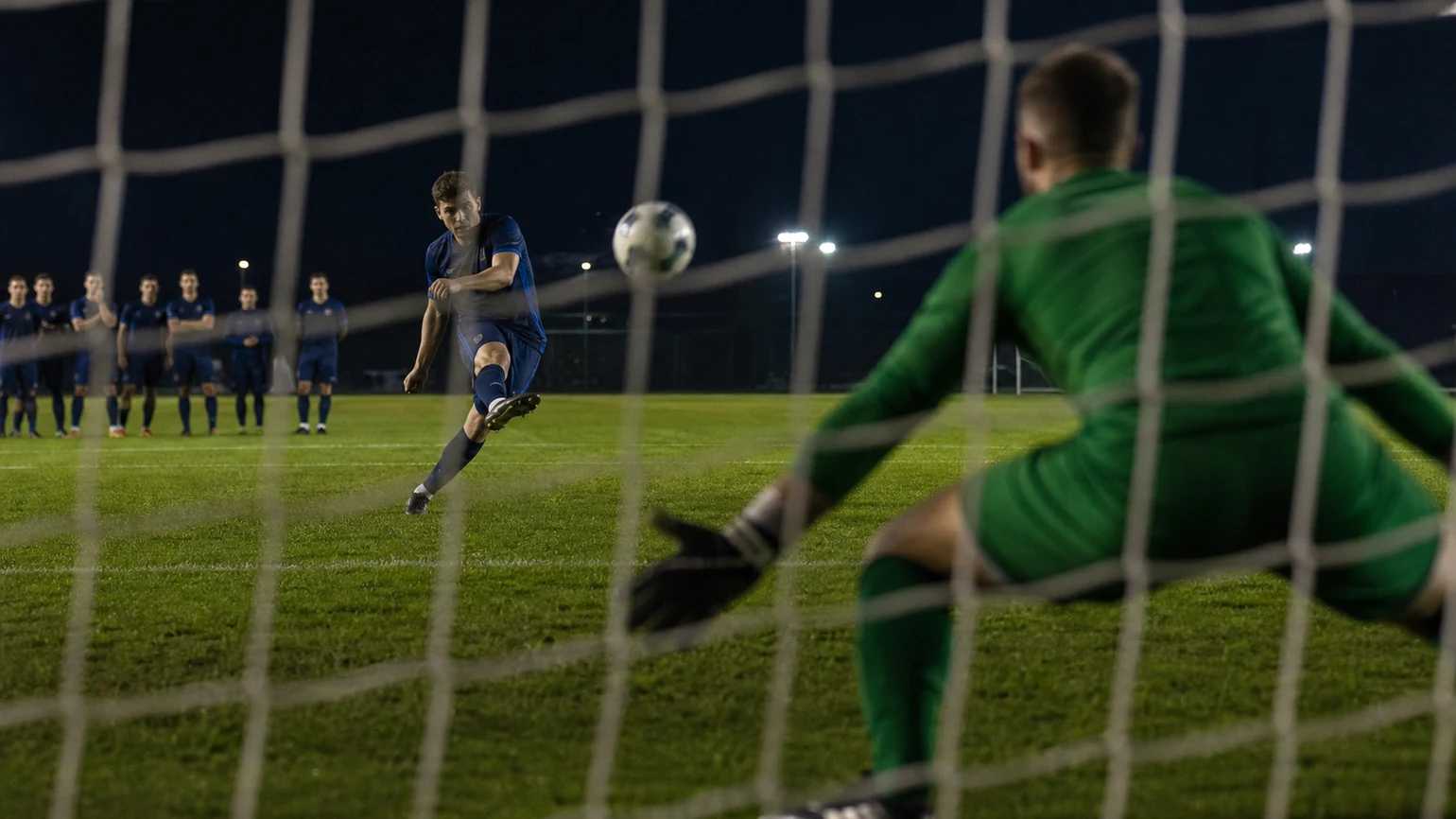 a striker's body shape set before a dropping ball near the penalty spot, keeper blurred beyond