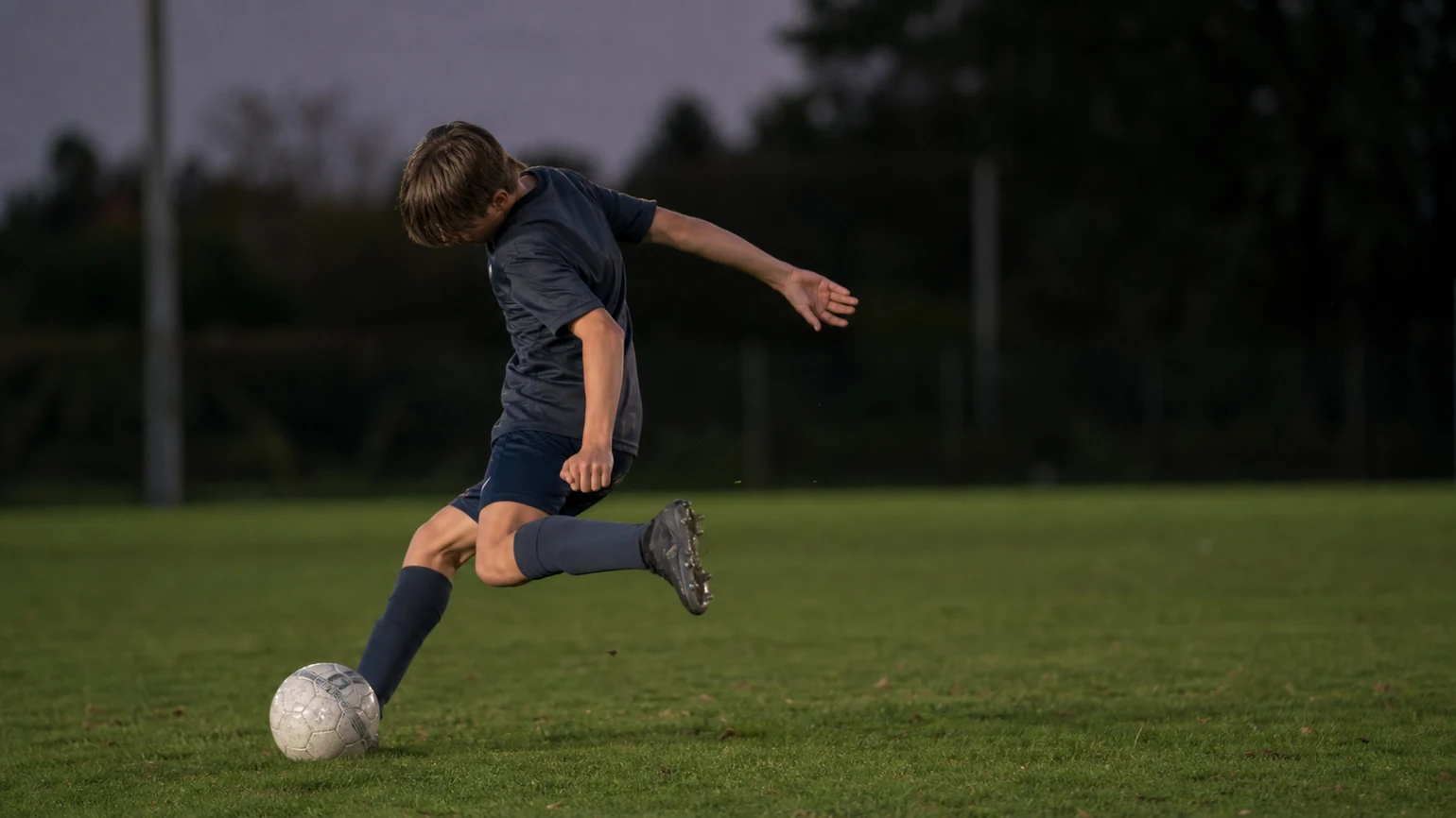 GENERATE — ONE 16:9 ultra-realistic documentary football photograph, low side angle at grass level: an anonymous youth player frozen at...